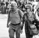 An elderly couple walks hand in hand through a busy city street.