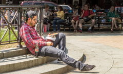 A man in a red plaid shirt sits on steps, reading a book in a lively park.