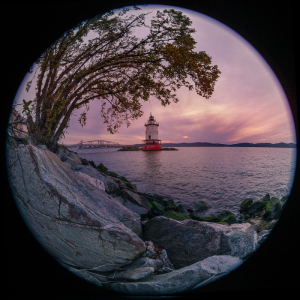 A lighthouse stands tall by the water, framed by trees and rocks, under a colorful sky.