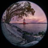 A lighthouse stands tall by the water, framed by trees and rocks, under a colorful sky.