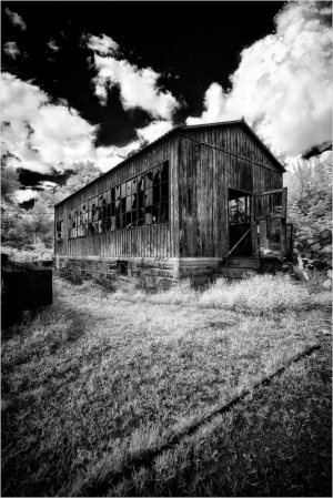 A weathered wooden building stands against a dramatic sky, windows shattered and overgrown grass surrounding it.