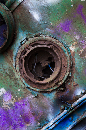 A rusty, colorful metal surface with a circular opening and blue wires hanging out. The vibrant decay tells tales of los