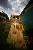 A rusted yellow bulldozer looms in a forgotten graveyard of machinery, overgrown with weeds.