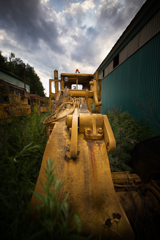 A rusted yellow bulldozer looms in a forgotten graveyard of machinery, overgrown with weeds.