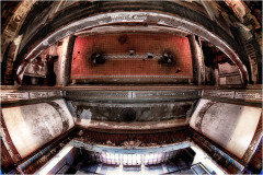 Interior of a decaying theater, with a wide view of an empty stage and ornate ceiling details.